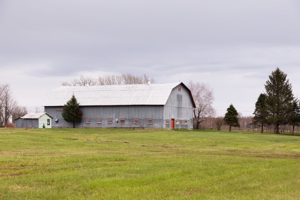 A large gray barn with a metal gambrel roof stands on a grassy field under a cloudy sky, surrounded by a few trees and bare branches in the background.