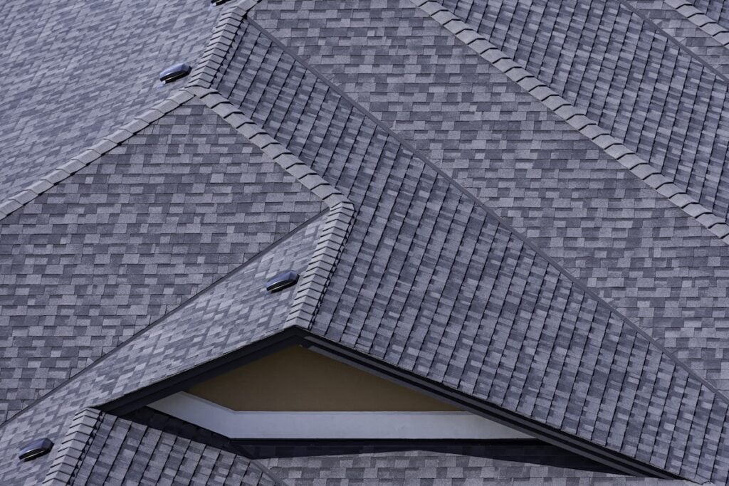 A close-up view of overlapping grey architectural shingles on rooftops, featuring repeating geometric patterns and textures with angled surfaces and clean lines.