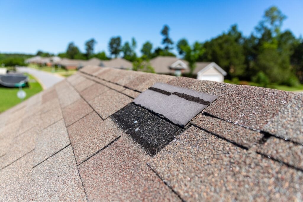 Close-up of a house roof with missing shingles on roof, highlighting one damaged or absent shingle that exposes the black underlayment. Neighboring houses and trees appear blurred in the background.