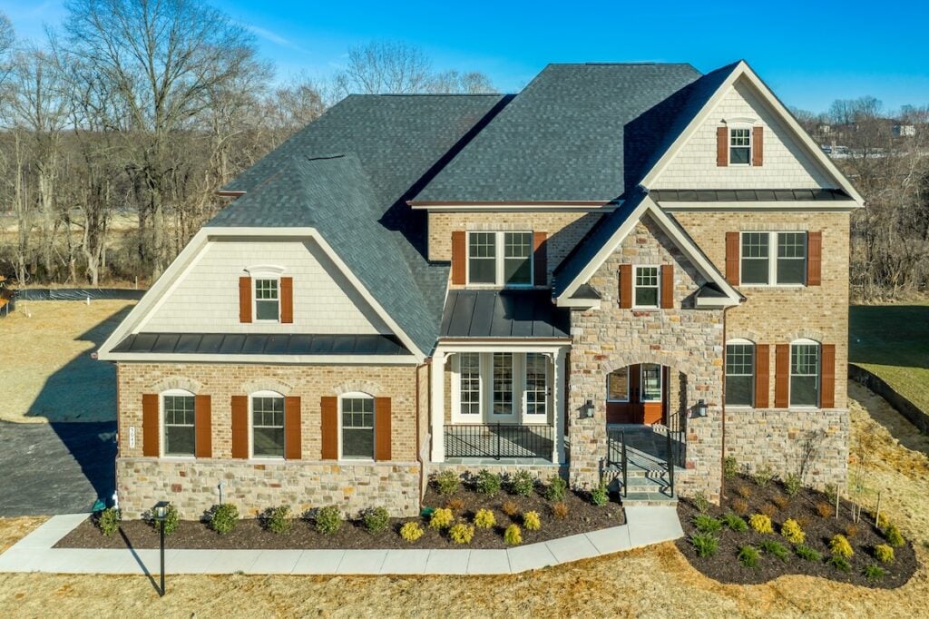 Two-story suburban house with a gambrel roof, brick and stone exterior, dark roof, white trim, and brown shutters. Multiple front windows, landscaped bushes, and a clear blue sky complete the scene.