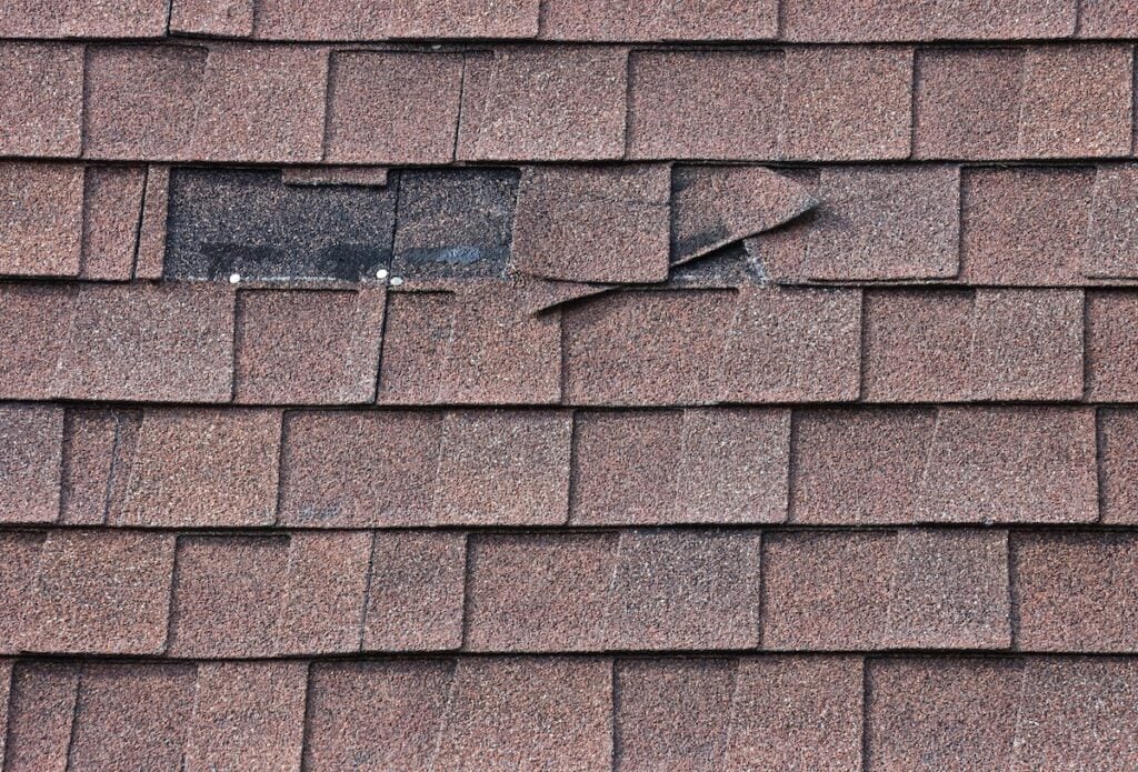 Several brown asphalt roof shingles are damaged, shifted out of place, and reveal black underlayment and some nails underneath. These missing shingles on roof areas could expose your home to leaks, though the surrounding shingles remain intact.