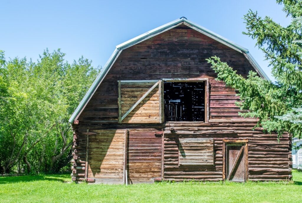 An old, weathered wooden barn with a gambrel roof and a partially open loft door stands on bright green grass, surrounded by leafy trees under a clear blue sky.