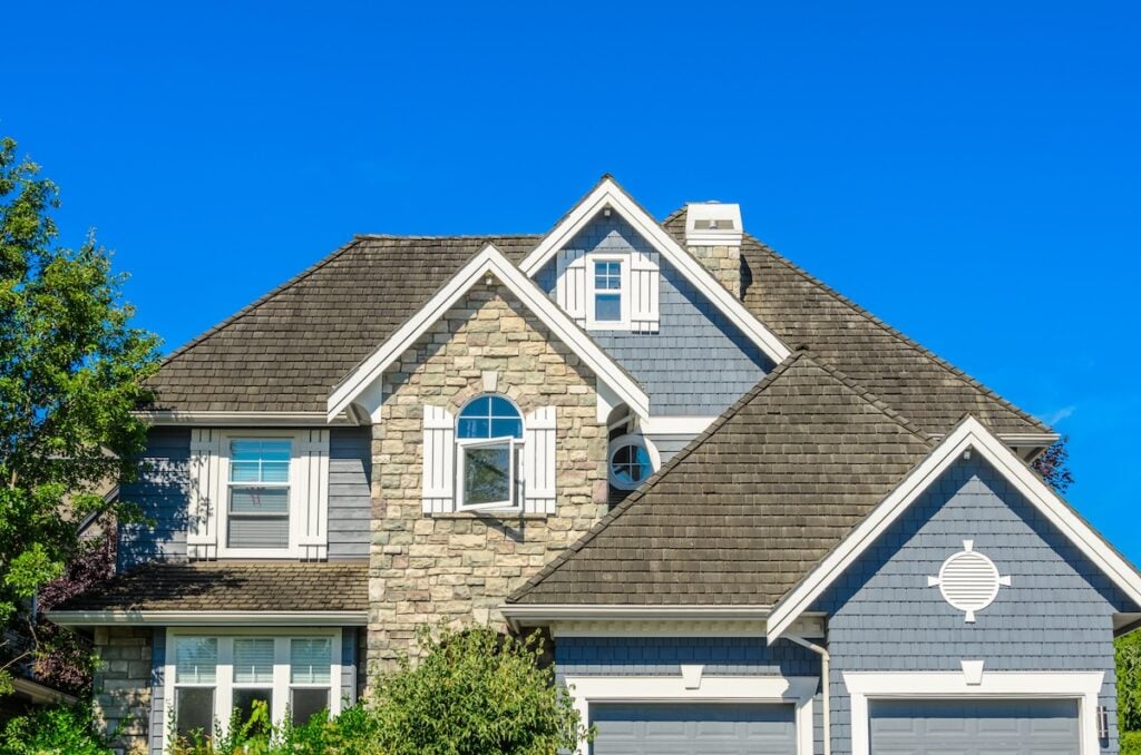 Two-story suburban house with gray siding and stonework, white shutters, multiple gables, and a dark shingle roof—ideal for those considering roof replacement cost—set against a clear blue sky with trees partially visible on the sides.