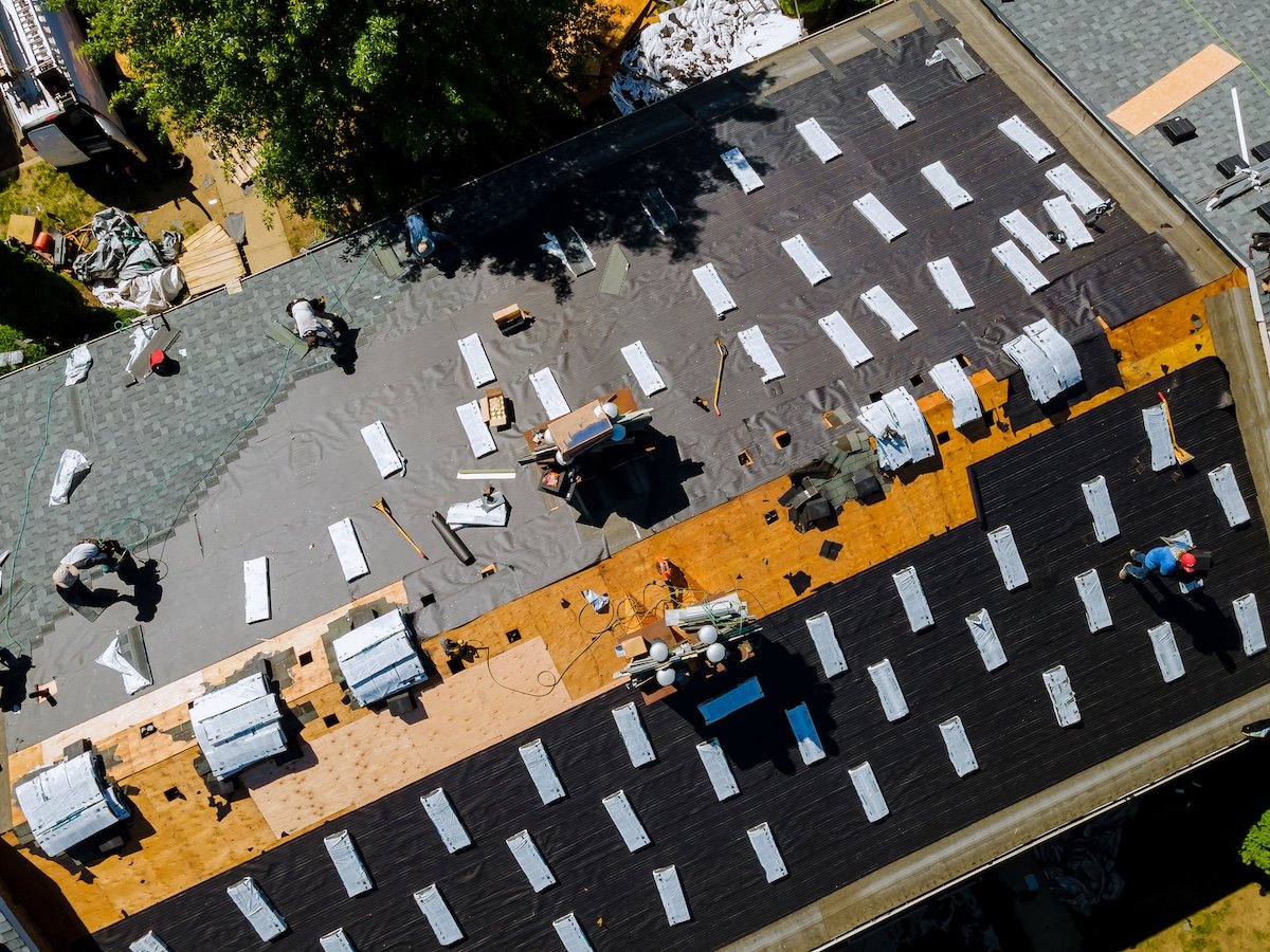 Aerial view of workers installing new roofing materials on a large house roof, with tools, tarps, and bundles of shingles spread across the surface, illustrating the scale and factors that can impact roof replacement cost.