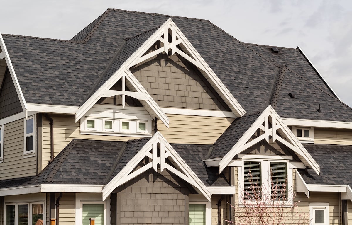 A close-up view of a house exterior featuring multiple steep, dark gray shingled rooflines, beige and gray siding, white trim, and several gables with decorative wooden accents.