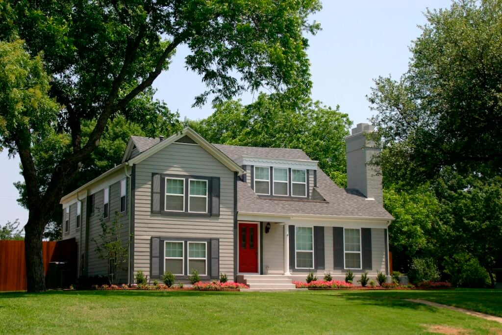 A two-story gray house with white trim and a red front door sits on a well-maintained lawn, surrounded by large green trees and landscaped flower beds under a clear, sunny sky.