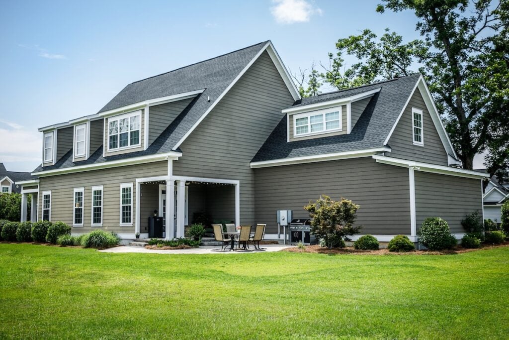 A modern two-story gray house with white trim, large windows, and a covered patio area. There is patio furniture and a grill outside, surrounded by a well-maintained lawn and landscaping.