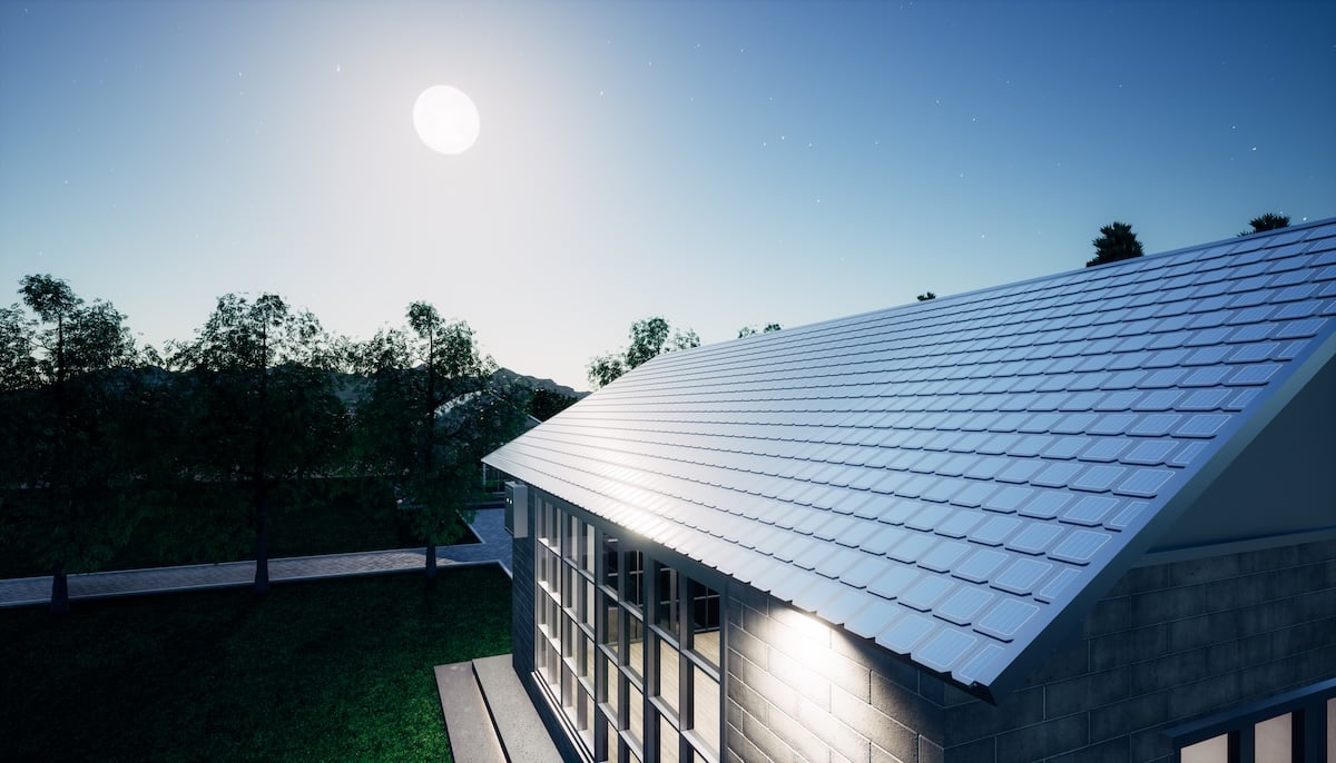 A modern house with a large metal roof and integrated solar shingles is illuminated by bright moonlight at night, surrounded by trees and a clear sky with visible stars.