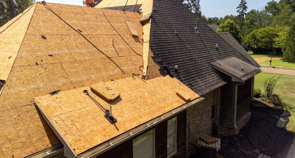 A house roof under construction shows new plywood sheathing alongside existing asphalt shingles, with tools and supplies on the roof—demonstrating stages of renovation that impact overall roof replacement cost.