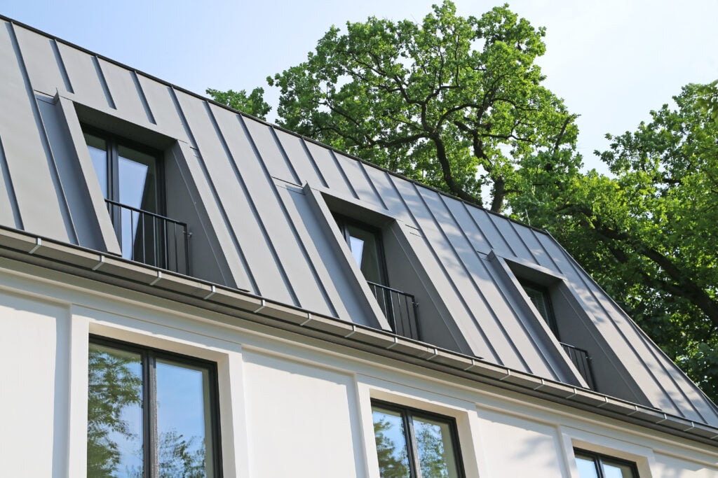 Modern house with a gray standing seam metal roof sloped and accented by three dormer windows, each with small black railings. The building is surrounded by green trees beneath a clear sky. standing seam metal roof