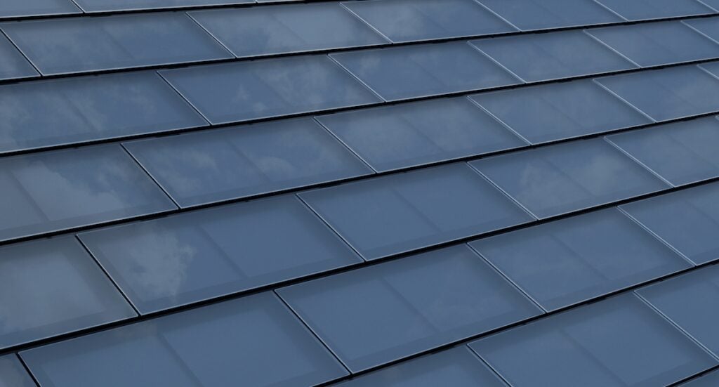 Close-up view of sleek, blue-toned solar shingles arranged in neat rows. The shingles have a smooth, glossy surface that reflects the sky and clouds above.