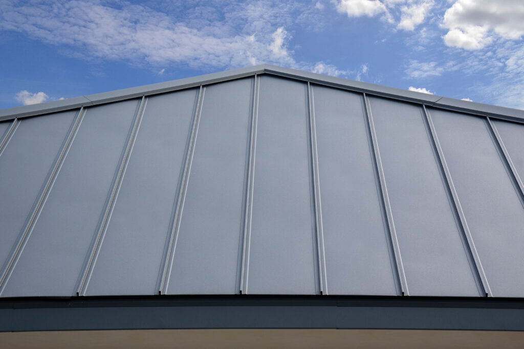 A close-up view of a modern standing seam metal roof with vertical seams, set against a partly cloudy blue sky. The geometric lines of the roof create a symmetrical pattern.
