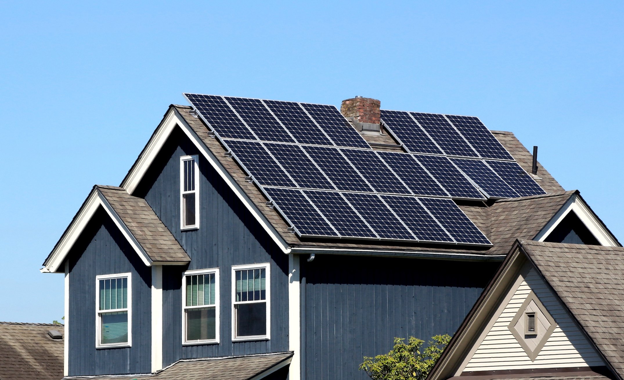 A dark blue house with multiple solar panels installed on its roof is pictured under a clear blue sky, prompting the question: how much energy does a solar panel produce? A neighboring house is visible in the foreground.