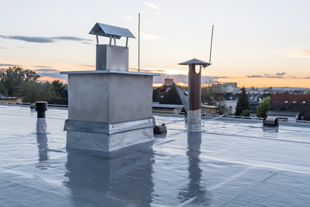A flat rooftop with vents and chimneys, covered in a reflective roofing membrane, overlooks a neighborhood at sunset beneath a clear sky dotted with scattered clouds.