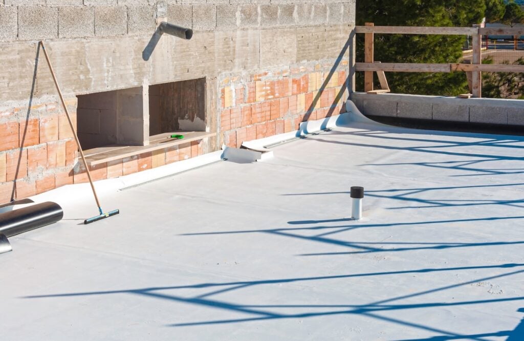 A flat rooftop under construction with a roofing membrane partially installed, tools and pipes scattered around, and unfinished brick and concrete walls enclosing the roof area. Professional workers waterproofing house construction site. Worker rolling membrane
