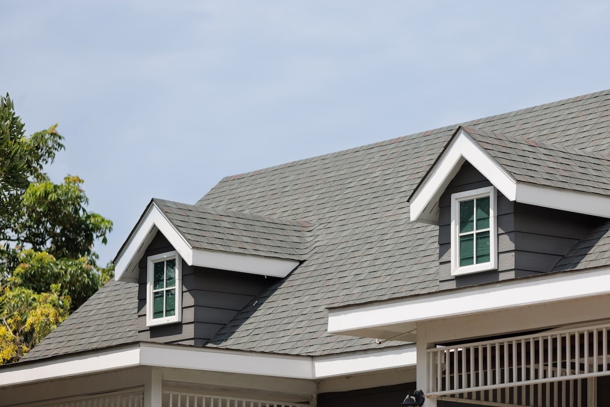 A close-up view of a house roof with gray shingles and two dormer windows, set against a partly cloudy sky and tree branches—a scene that highlights the classic appeal in the metal roof vs shingles debate.