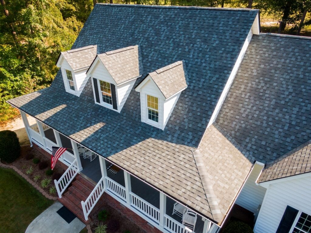 Aerial view of a house with gray shingle roofing, white siding, and three dormer windows—an ideal example when considering metal roof vs shingles. A front porch with a white railing and an American flag is surrounded by green trees and landscaping. metal roof vs shingles
