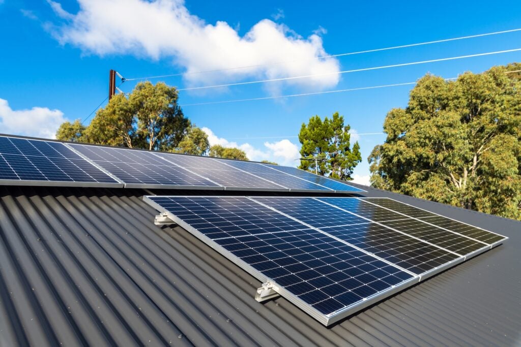 Solar panels on metal roof absorb sunlight atop a dark corrugated surface, framed by green trees and a blue sky with clouds in the background. Power lines stretch above the treetops, completing the scene.