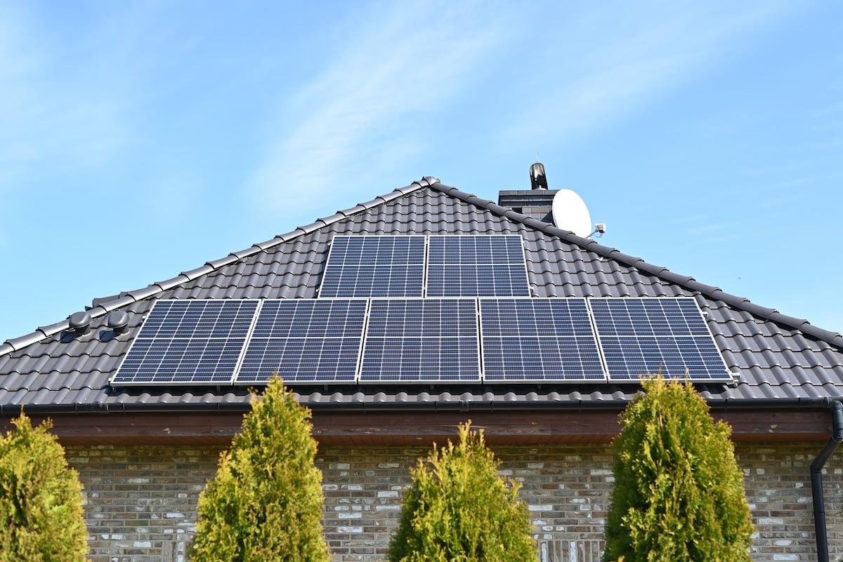 A house with a sloped dark tiled roof features multiple solar panels and solar battery storage on top. Green trees fill the foreground, while a clear blue sky completes the scene.