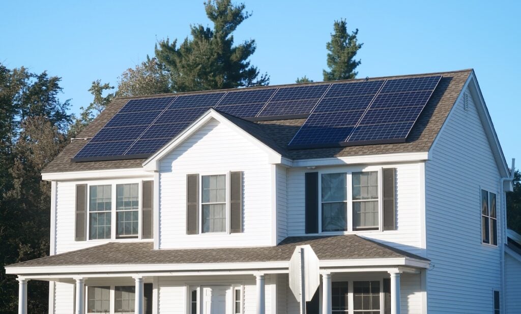 A white two-story house with dark shutters features several solar panels and a solar battery storage system on its roof, surrounded by trees under a clear blue sky. solar battery storage