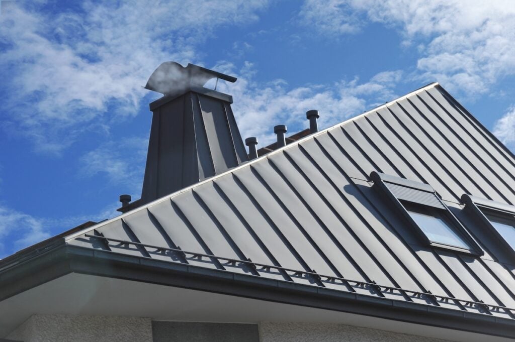 A modern metal roof with a steep pitch, featuring a chimney topped with a metal cap, several vent pipes, and a skylight window, highlights the advantages of metal roof vs shingles against a partly cloudy blue sky. metal roof vs shingles