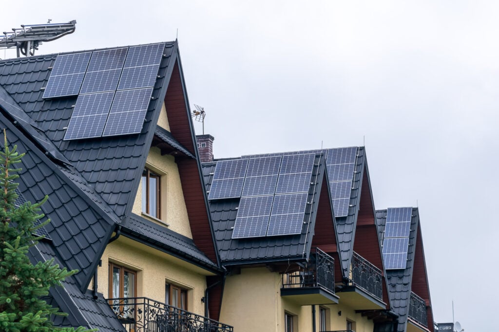 Row of houses with dark, sloped metal roofs, each fitted with solar panels on metal roof. The buildings have balconies with ornate railings, and the sky is overcast.