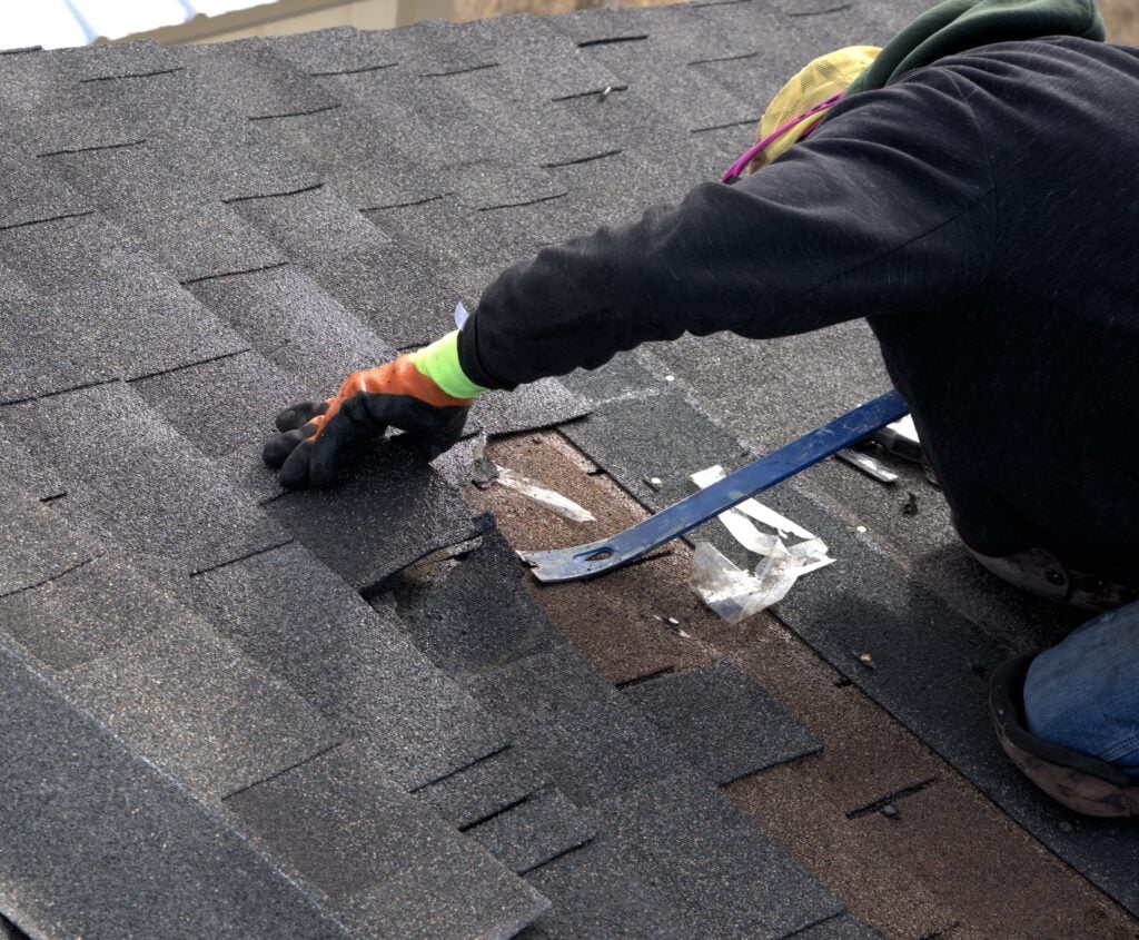 A person wearing gloves and a hat performs roof repair by using a pry bar to lift damaged asphalt shingles, preparing to replace or fix them. roof repair