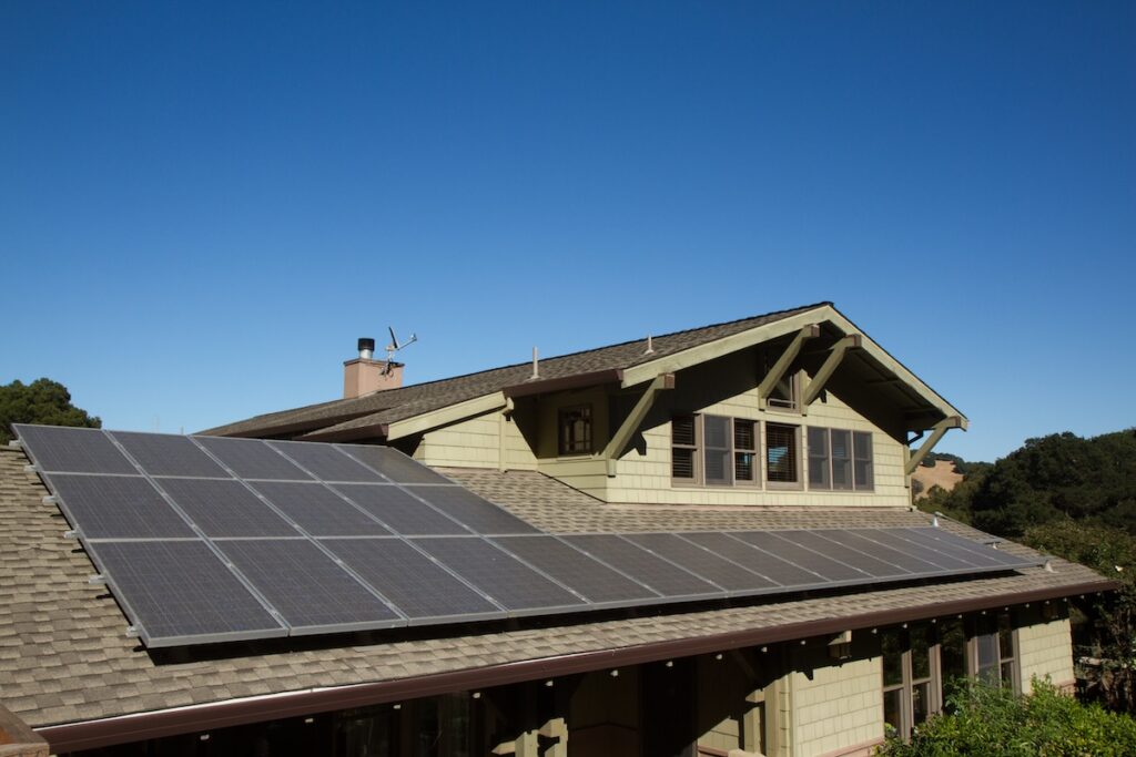 Solar panels installed on the roof of a green house beneath a clear blue sky, demonstrating how many solar panels to power a house, with trees and hills visible in the background. how many solar panels to power a house