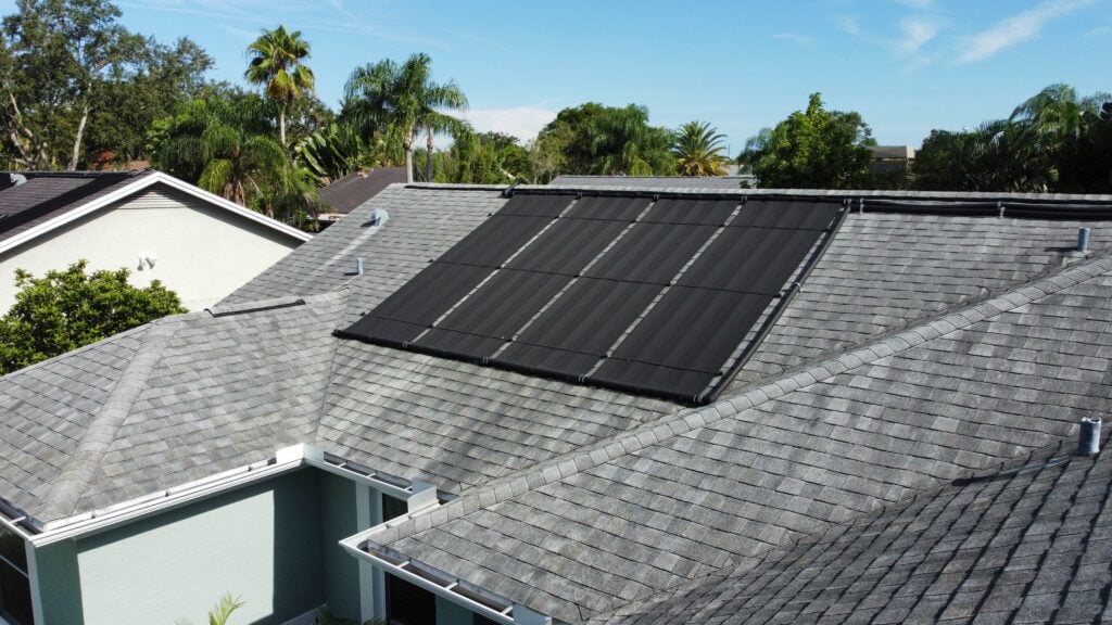 A rooftop with gray shingles features several black solar panels installed on one section, offering a glimpse of how many solar panels to power a house. Surrounding homes, trees, and a clear blue sky are visible in the background. how many solar panels to power a house