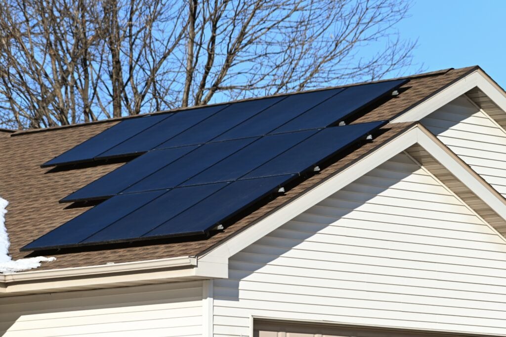 Solar panels installed on the roof of a suburban house with beige siding demonstrate how many solar panels to power a house; bare tree branches and a blue sky are visible in the background. how many solar panels to power a house