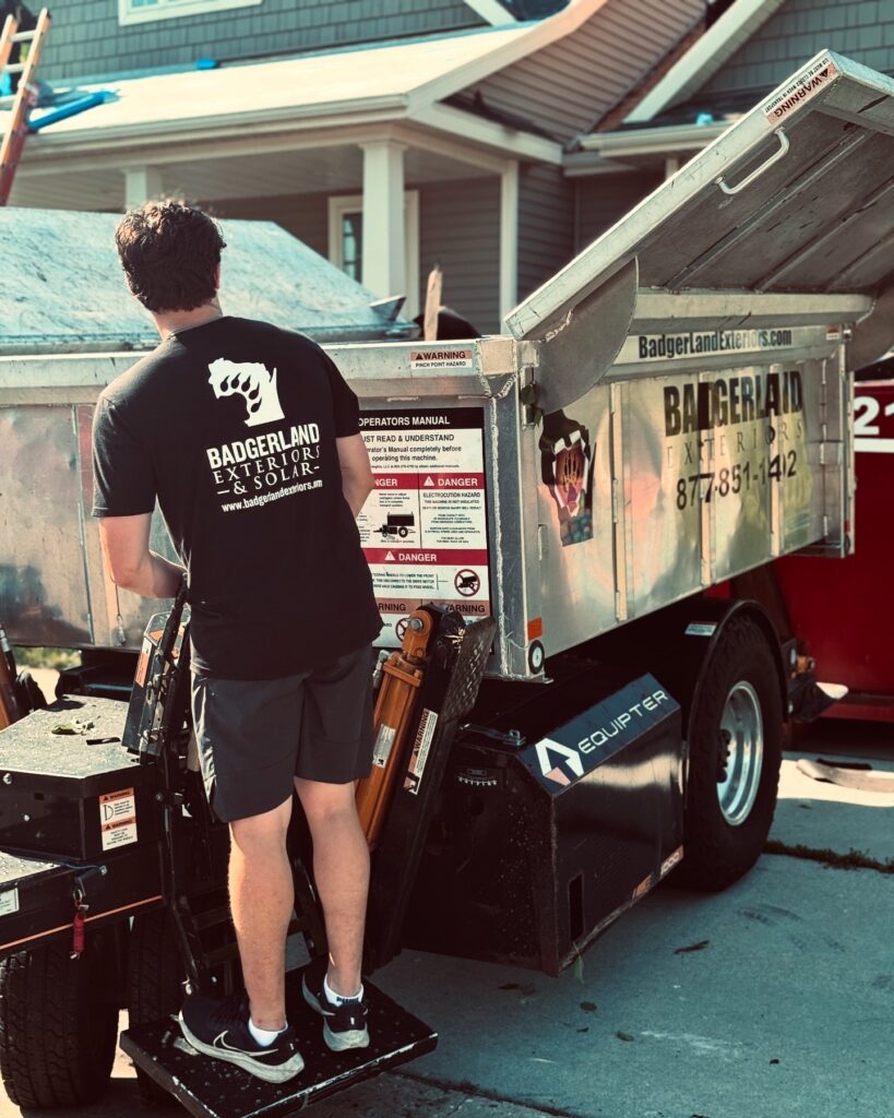A man wearing a "Badgerland Exterior & Solar" t-shirt operates a hydraulic lift next to a metal trailer labeled "Badgerland Exteriors" outside a residential house on a sunny day.