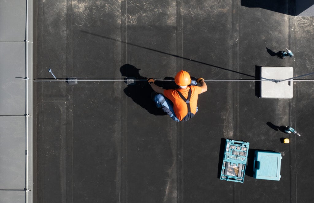 A worker in an orange vest and helmet kneels on a flat black roof, using tools from an open blue toolbox nearby, as they perform commercial roof maintenance along a safety line. The photo is taken from directly above.