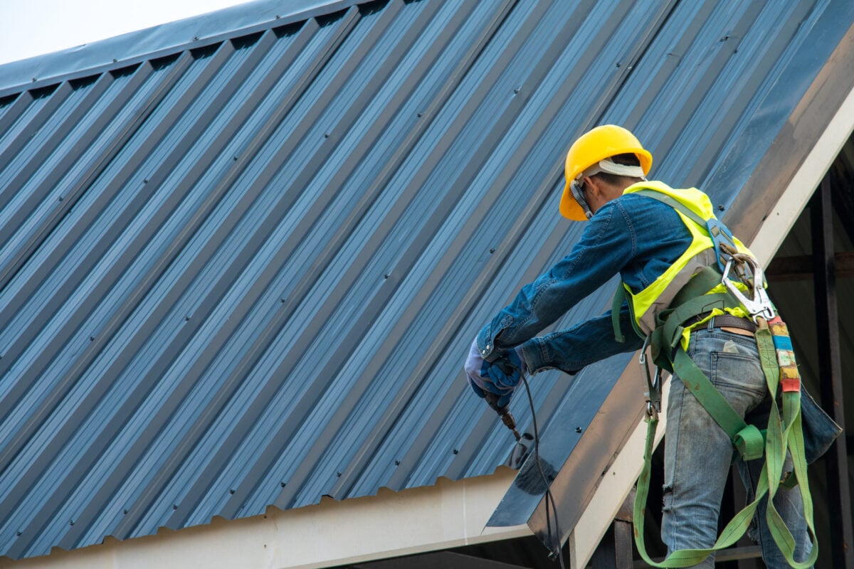A construction worker wearing a yellow hard hat, safety harness, and reflective vest installs metal roofing panels as part of commercial roof maintenance. The worker handles tools while standing near the edge of the roof.
