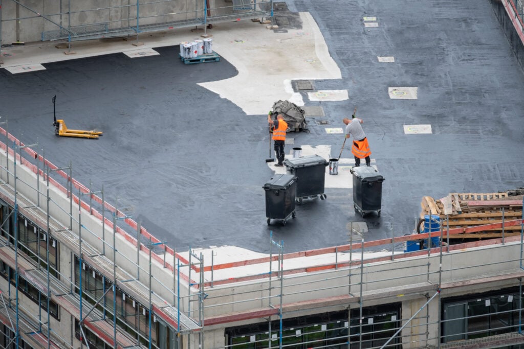 Two construction workers in orange vests perform commercial roof maintenance on a flat rooftop, applying black material. Scaffolding surrounds the building, with equipment and materials scattered nearby.