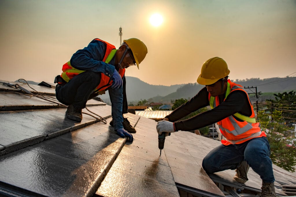 Two construction workers wearing safety vests and helmets install roofing panels with tools on a sloped roof at sunset, demonstrating commercial roof maintenance amid hills and trees in the background. commercial roof maintenance
