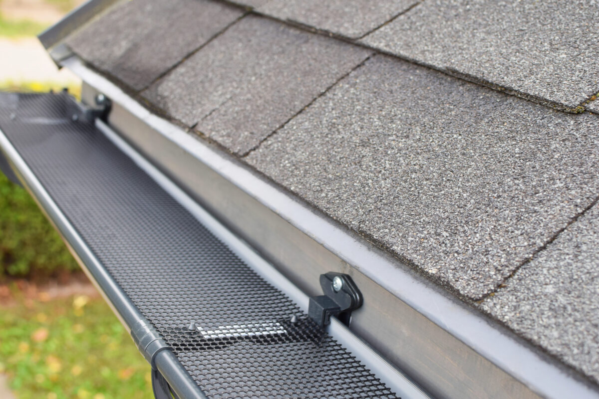 Close-up view of a house roof with asphalt shingles and a black mesh gutter guard installed along the edge, showcasing how to install gutter guards to prevent debris from entering the rain gutter.