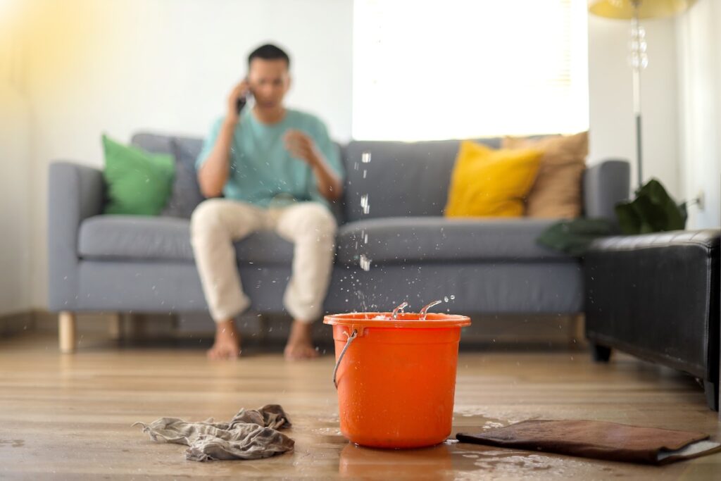 metal roof leak repair A man sits on a couch talking on the phone while water drips from the ceiling into an orange bucket, wet rags and a towel scattered nearby—signs that a metal roof leak repair is urgently needed.