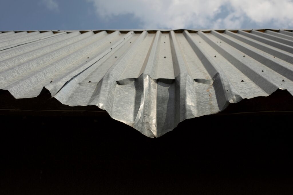 A close-up view of a metal roof hail damage corrugated metal roof with a large dent or bend at the edge, set against a partly cloudy sky.