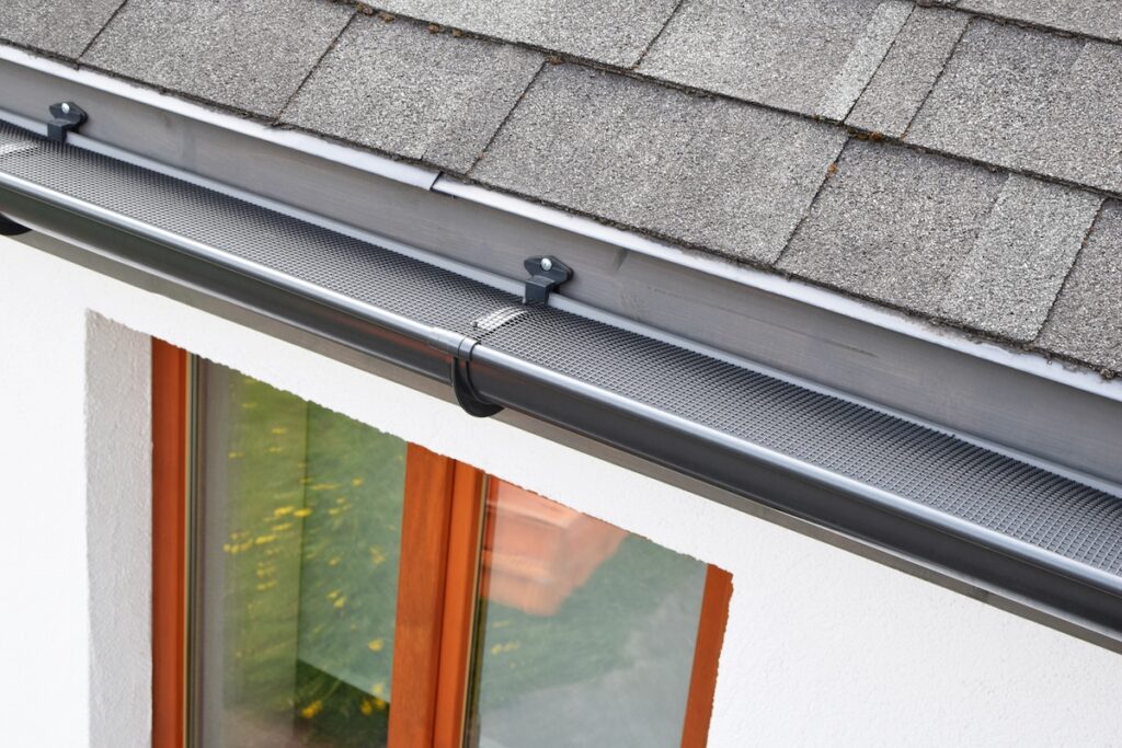 A close-up view of a house roof with gray shingles, a black gutter fitted with a metal mesh guard (ideal when asking, "are gutter guards worth it?"), and a window with a wooden frame below. The white exterior wall is also visible. are gutter guards worth it