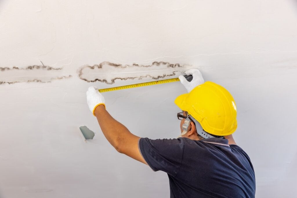 metal roof leak repair A worker in a yellow hard hat and gloves is measuring a water-stained crack on a white ceiling with a measuring tape, inspecting for possible metal roof leak repair. The worker is facing away from the camera.