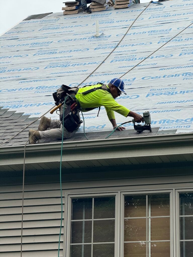 A construction worker in a safety harness installs shingles on a roof using a nail gun. He is wearing a bright yellow shirt, work gloves, and a cap. Roofing Madison materials and tools are visible around him.