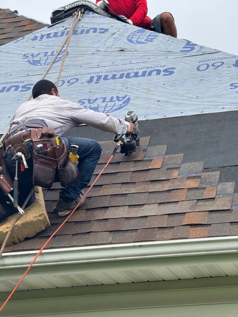 Two workers from Roofing Madison install new shingles; one uses a nail gun while wearing a tool belt, and the other kneels on roofing underlayment above, both secured with harnesses.
