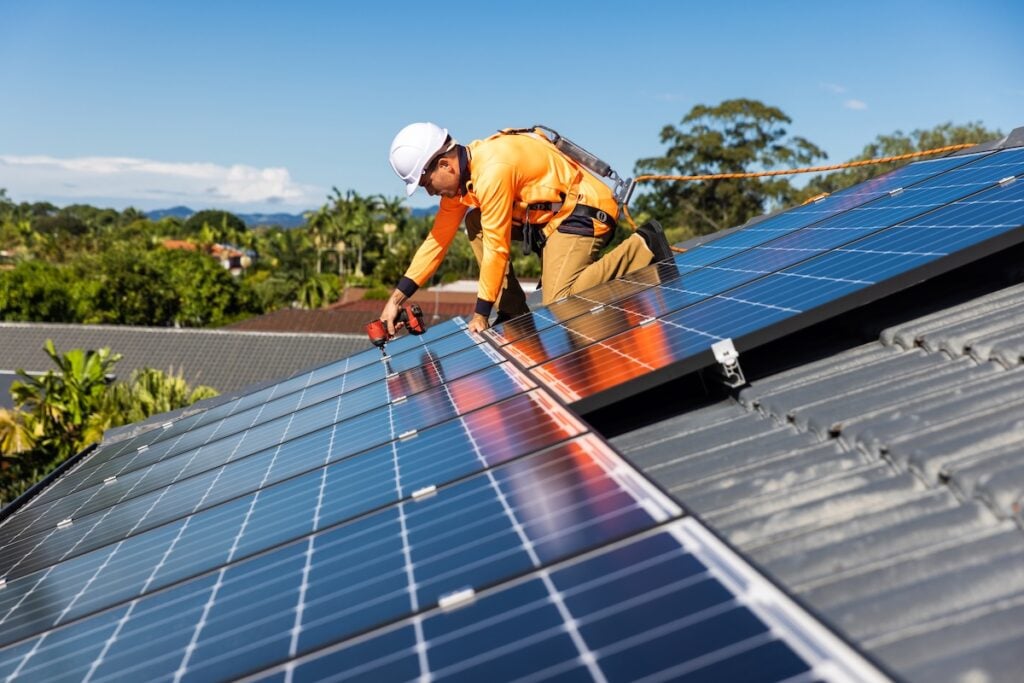A worker in a safety harness and helmet installs solar panels on a residential rooftop under a clear blue sky, reminding homeowners of the importance of preparing questions to ask solar companies before starting their own installations.