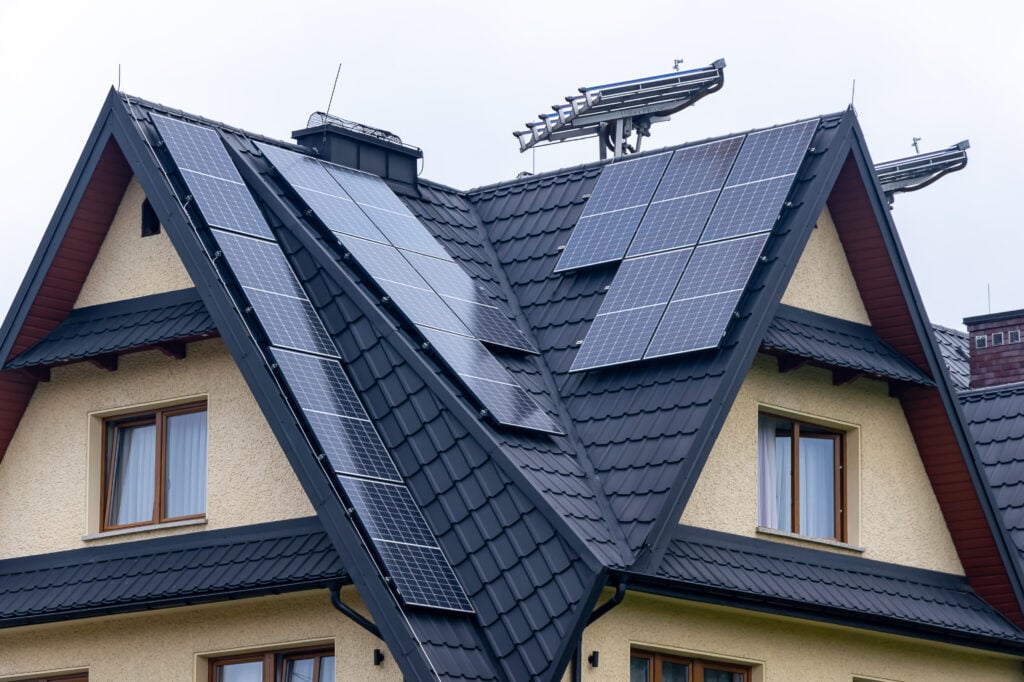 A house with beige walls and a dark tiled roof has multiple solar panels installed on its sloped surfaces. Antennas are also mounted on the roof. When considering solar, remember to research questions to ask solar companies for peace of mind. The sky is overcast. questions to ask solar companies