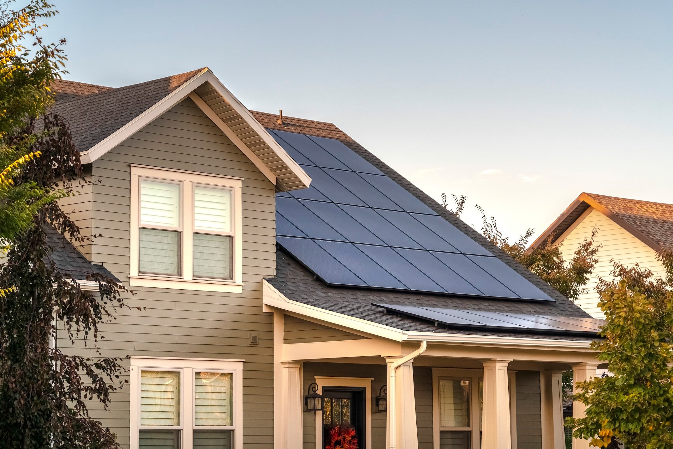 A modern two-story house with gray siding features multiple solar panels on its roof, surrounded by trees and bushes under a clear blue sky—perfect inspiration if you have questions to ask solar companies about home installations.