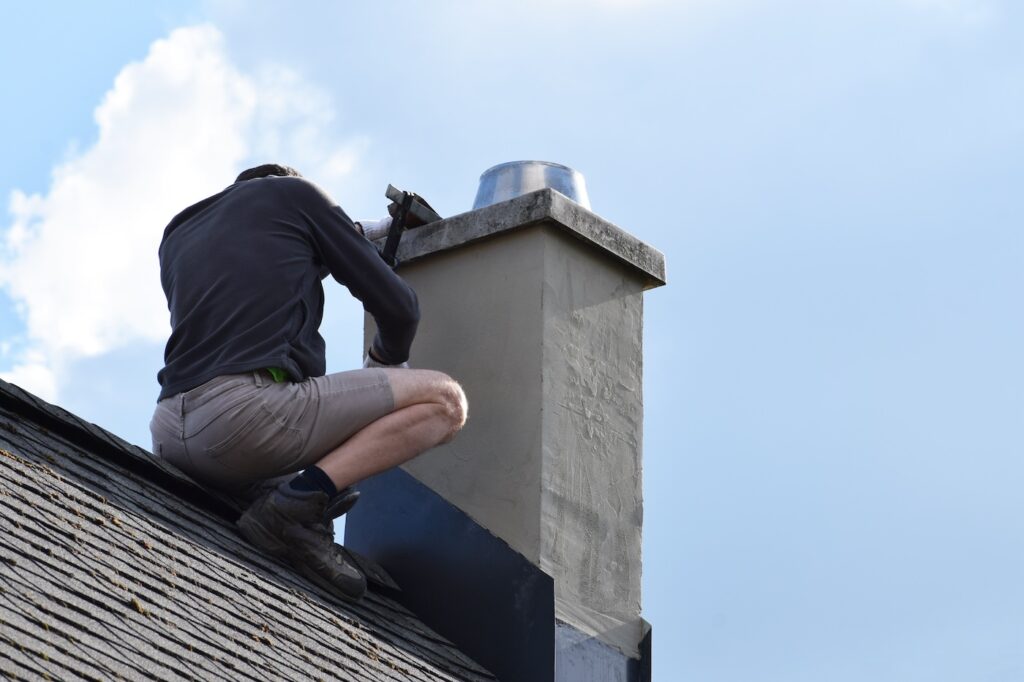 A person wearing shorts and a long-sleeve top is kneeling on a sloped roof, working on a chimney with a tool, under a partly cloudy sky.