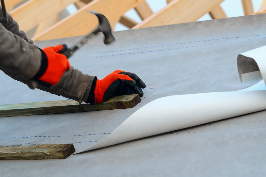 A person wearing orange gloves uses a hammer to secure a wooden beam on a roof covered with a large sheet of roofing material, with more roofing paper being unrolled nearby.