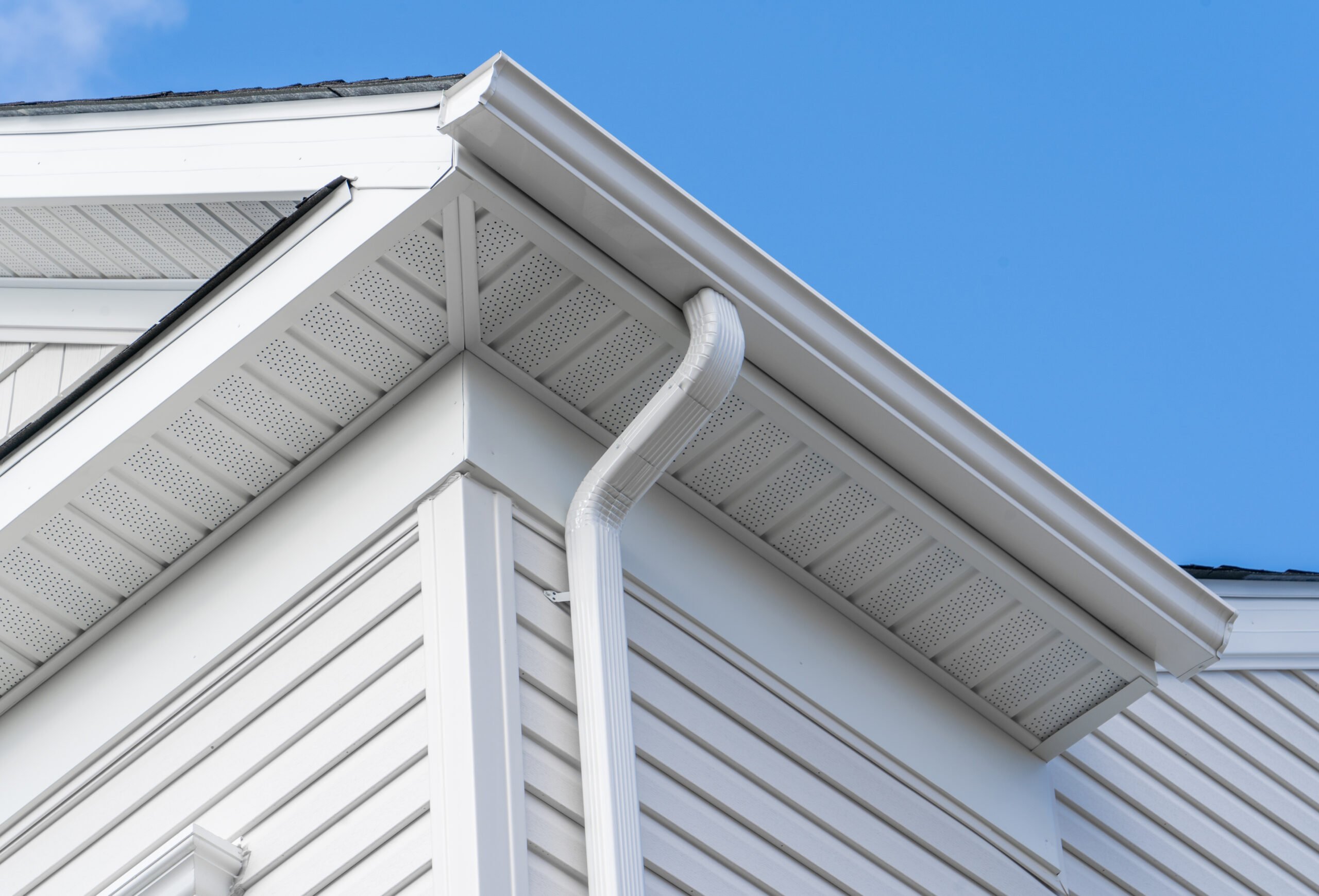 A close-up view of a house's white vinyl siding, soffit, fascia, and a rain gutter system with a downspout against a clear blue sky.