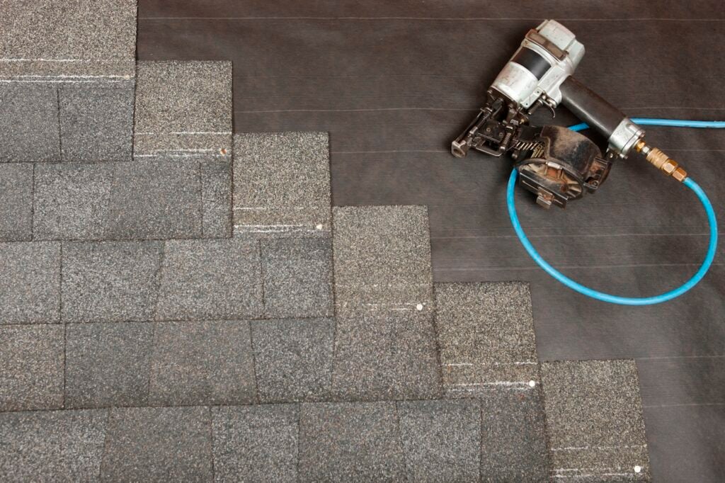 Close-up of gray asphalt roof shingles being installed in a staggered pattern on a roof, next to a pneumatic nail gun with a blue air hose lying on black underlayment.
