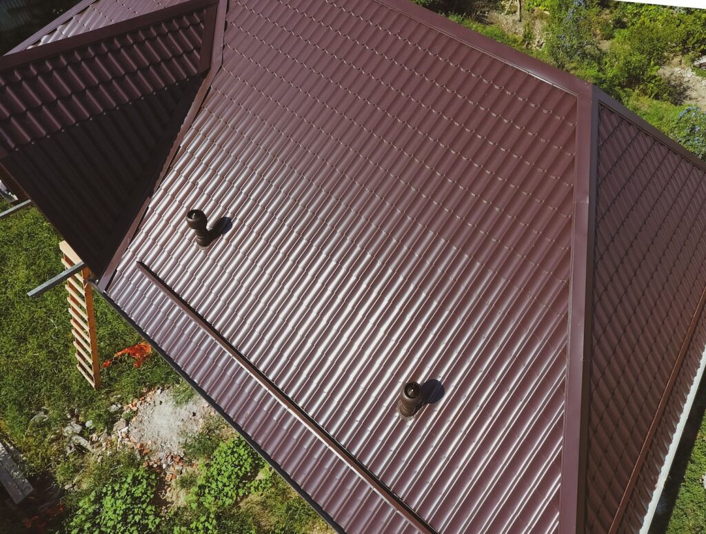 Aerial view of a brown metal roof with two vent pipes, surrounded by green grass and plants. The roof features ridged panels and an angular design.